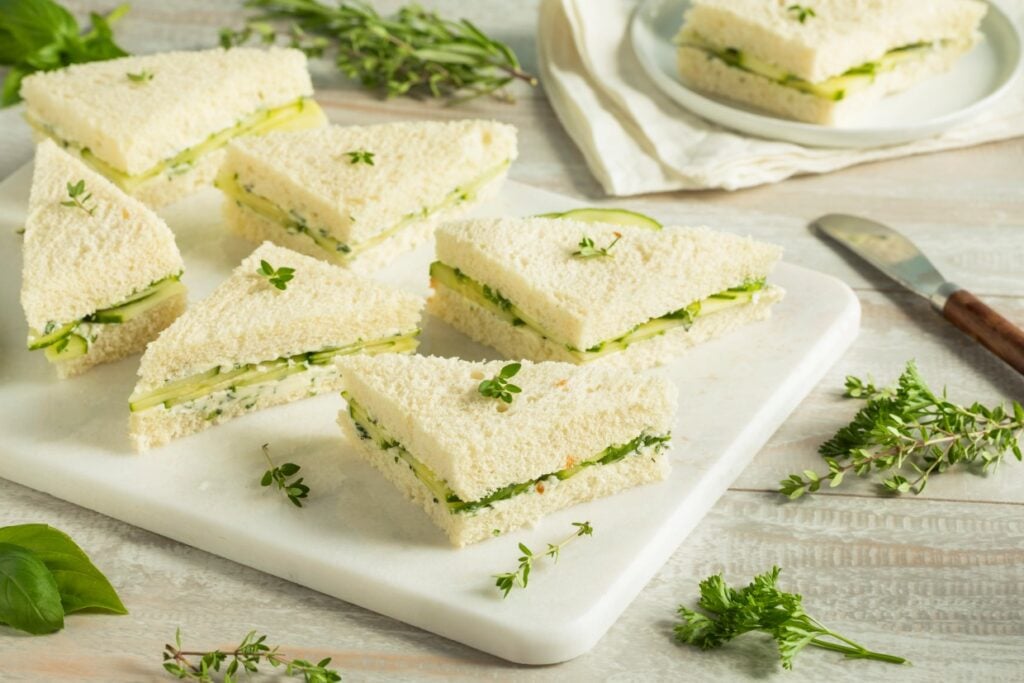 Triangular cucumber sandwiches with cream cheese and herbs are neatly arranged on a white cutting board. Fresh herbs are scattered around, and a knife rests nearby. There's a stack of small plates in the background.