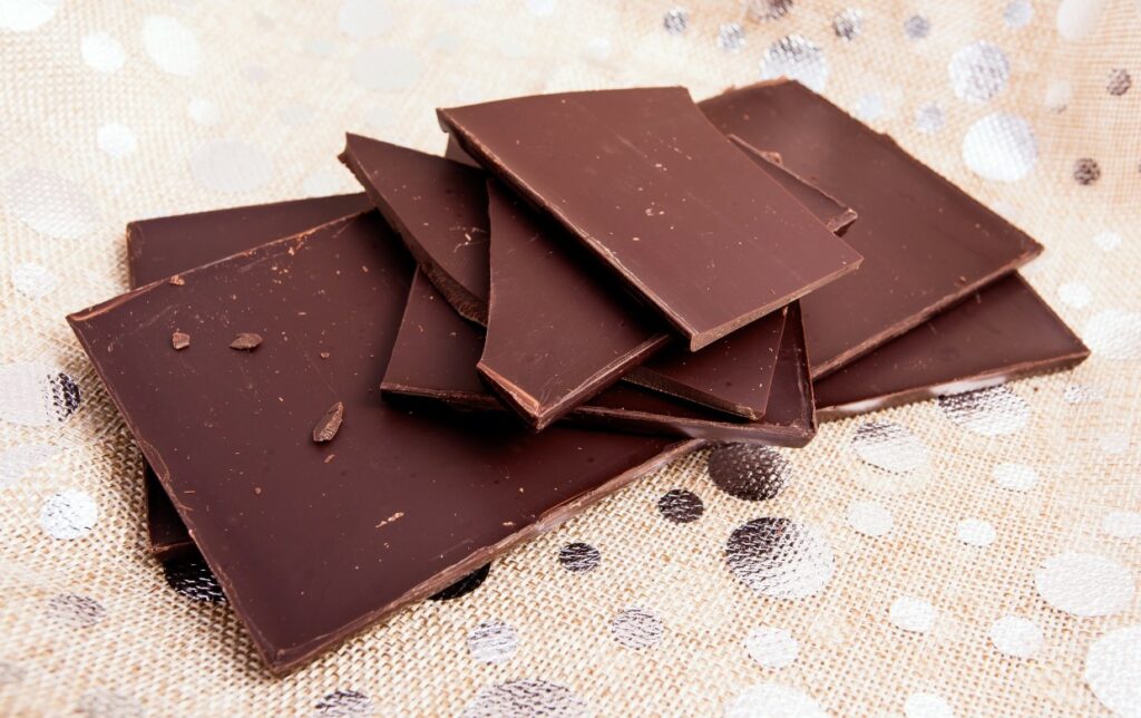 Several pieces of dark chocolate are stacked on a textured, metallic surface with a polka dot pattern. The chocolate appears glossy and smooth with a few irregular edges.