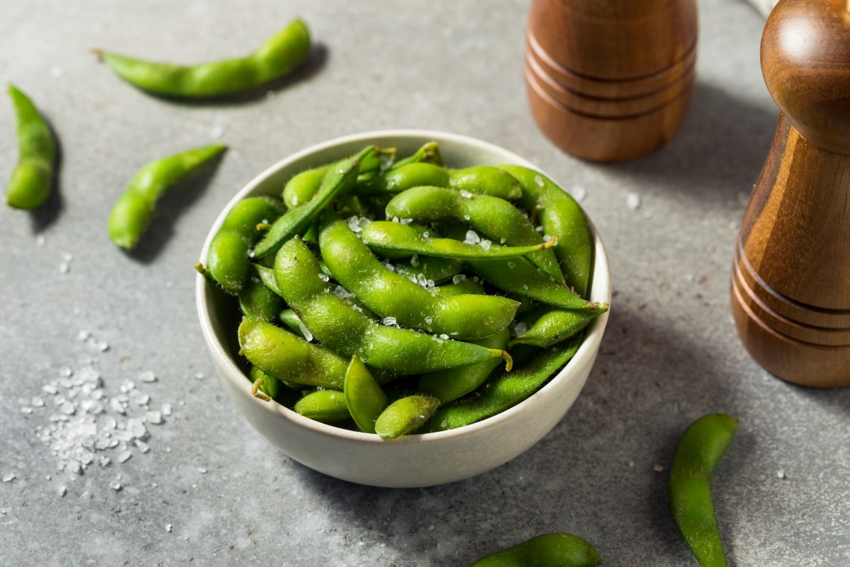 A bowl of salted edamame with pods, surrounded by scattered salt crystals on a gray surface. Wooden salt and pepper grinders are visible on either side of the bowl.