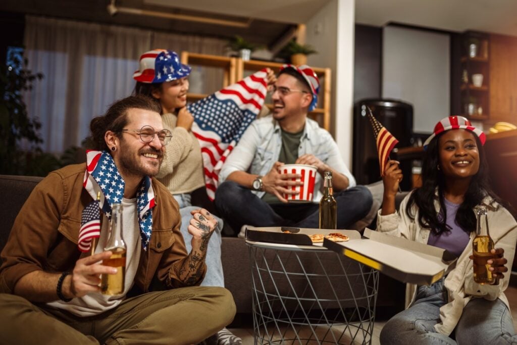 A group of four friends wearing American flag-themed attire sit together in a cozy room. They hold small American flags and snacks, including popcorn and pizza, while smiling and enjoying drinks. The atmosphere is festive and relaxed.