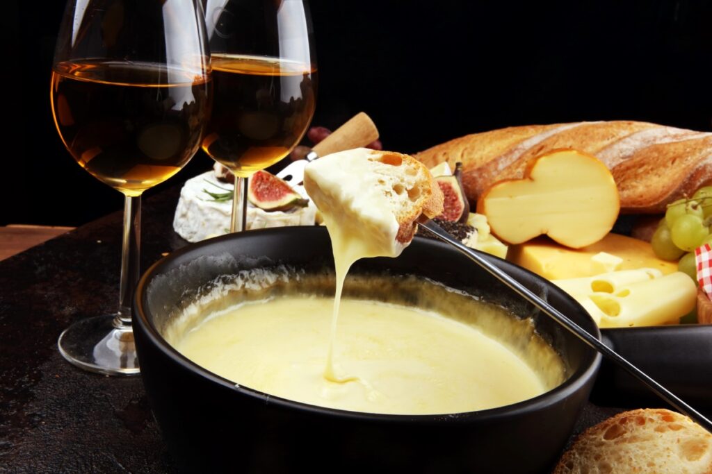 A black bowl with melted cheese fondue, a piece of bread being dipped. In the background, there are assorted bread, sliced cheese, figs, and grapes. Two glasses of white wine are visible, creating an inviting, cozy atmosphere.