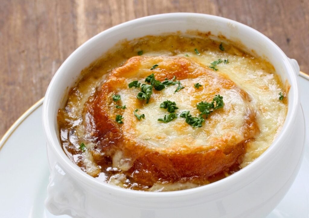 A bowl of French onion soup topped with a slice of toasted bread and melted cheese, garnished with chopped parsley. The soup is served in a white ceramic bowl on a wooden table.