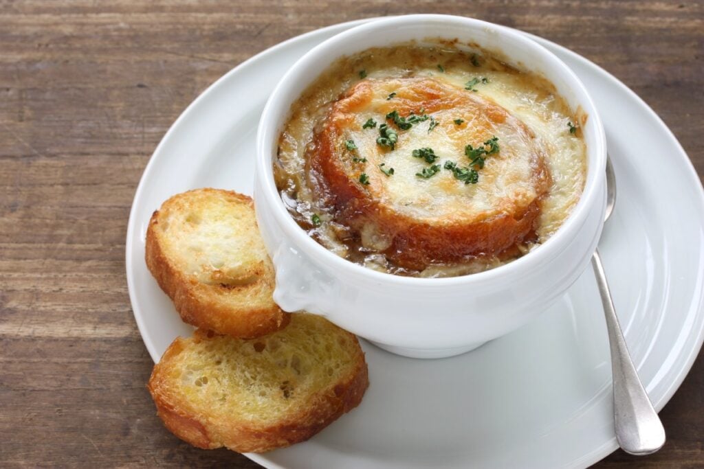 A bowl of French onion soup topped with melted cheese and herbs, served on a white plate. Two slices of toasted baguette are placed beside the bowl. A spoon rests on the plate. The background is a wooden table.