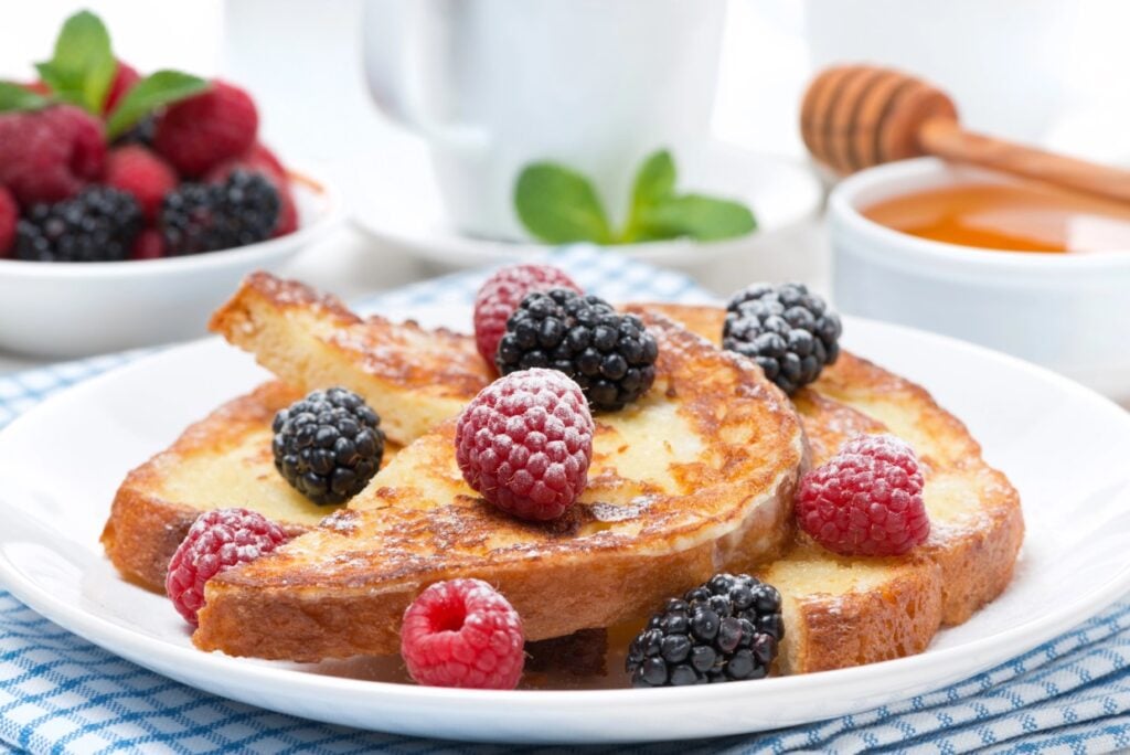 A plate of French toast topped with fresh raspberries and blackberries. Nearby, a bowl filled with raspberries and a honey jar with a dipper are visible. White cups and a mint garnish complete the breakfast setting on a checkered cloth.