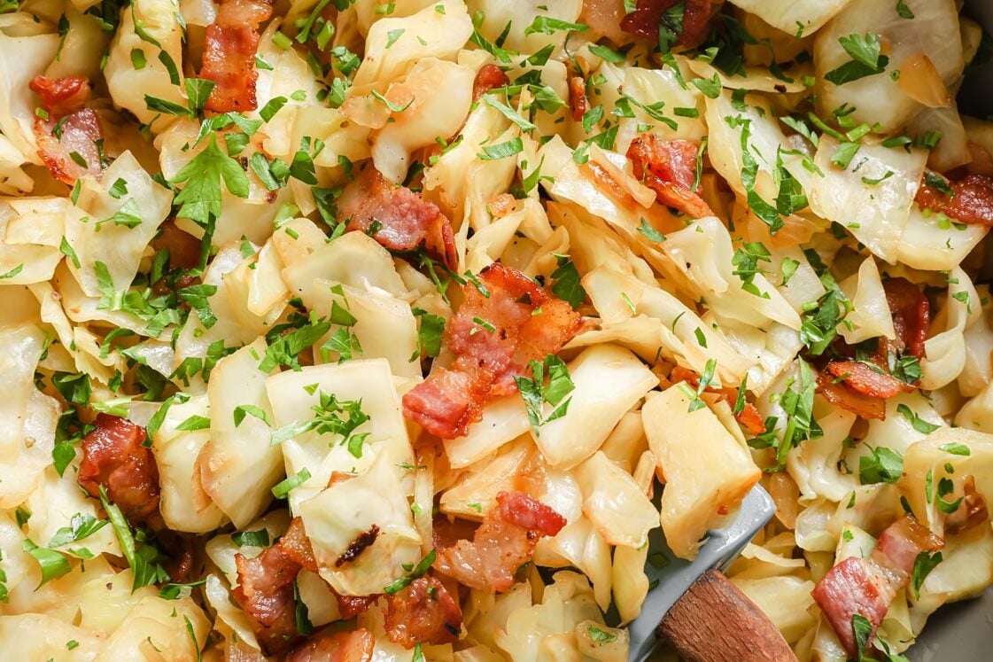 A pan full of bacon and cabbage with a wooden spoon.