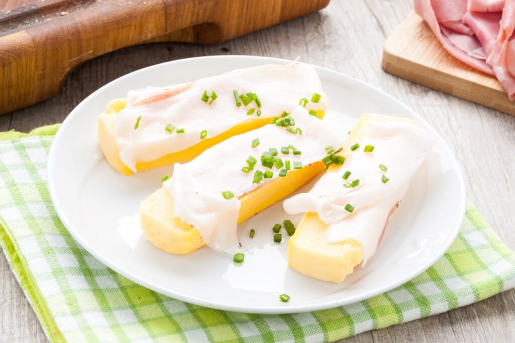 A plate with three slices of cornbread topped with thin slices of white cheese and garnished with chopped chives. The plate sits on a green checkered cloth, with sliced ham on a wooden board in the background.