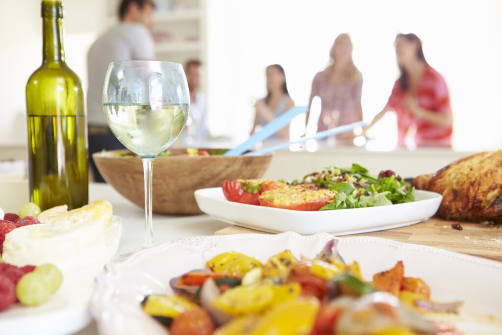 A table with a variety of dishes, including salad, grilled vegetables, and roasted chicken. A bottle of wine and a glass of white wine are in the foreground. In the background, blurred figures are socializing.