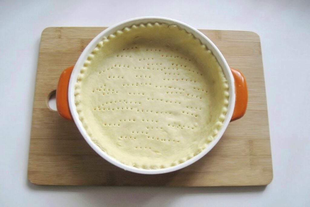 A round, unbaked pie crust in a white dish with orange handles, on a wooden cutting board. The crust is evenly pricked with a fork across the surface.