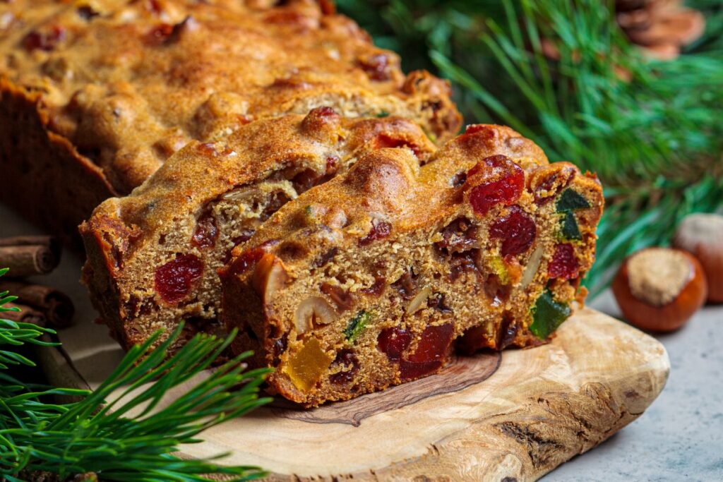 Close-up of a sliced fruitcake on a wooden board. The cake is dense with visible chunks of dried fruits and nuts. Pine branches and hazelnuts decorate the background, creating a festive atmosphere.