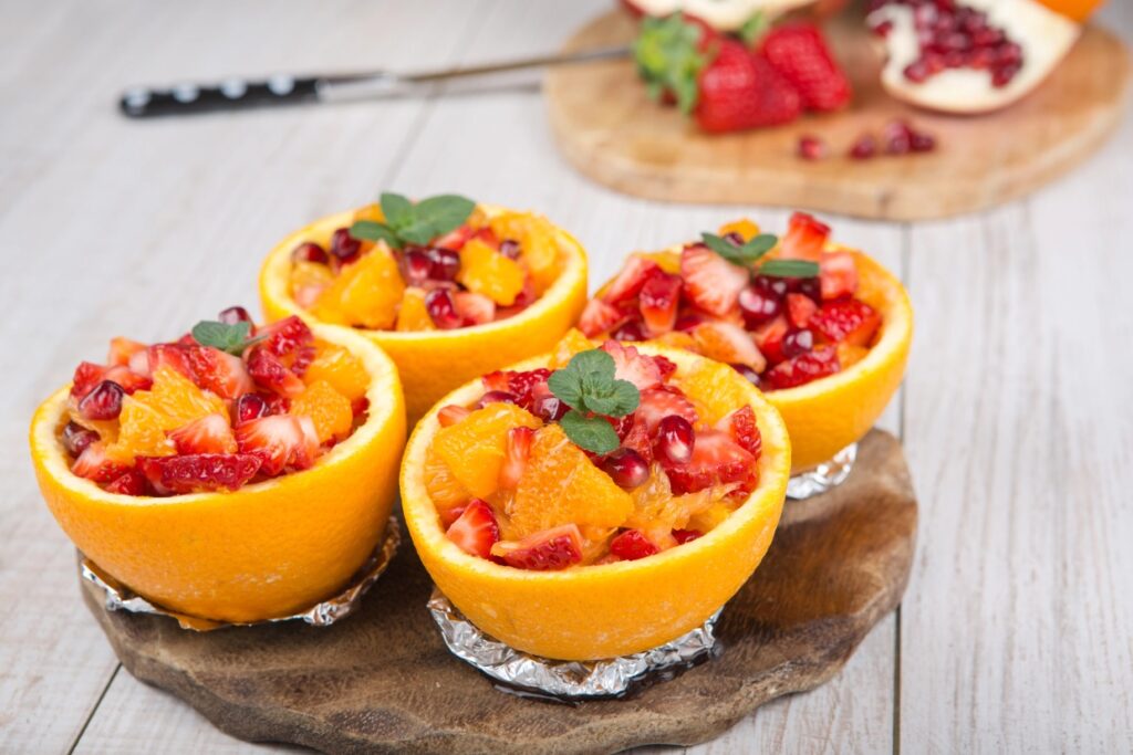 Four orange halves filled with a colorful fruit salad consisting of strawberries, oranges, and pomegranate seeds. The oranges are placed on a wooden board with a backdrop of strawberries and a sliced pomegranate.