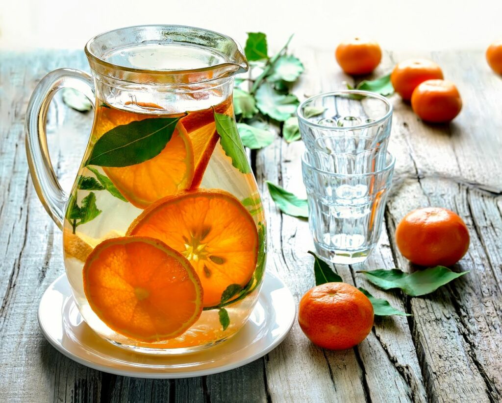 A glass pitcher filled with citrus-infused water, featuring slices of oranges and a few green leaves. It's placed on a white plate. Beside it are stacked glasses and scattered whole oranges with leaves on a rustic wooden table.