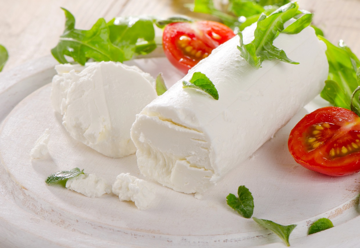 A log of soft white cheese on a wooden board, garnished with fresh arugula leaves and halved cherry tomatoes.