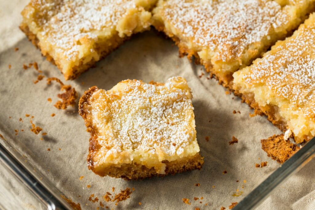 A close-up of gooey butter cake pieces in a baking dish. One piece has been separated from the rest, showing a moist, crumbly texture. The cake is dusted with powdered sugar and rests on a beige cloth.