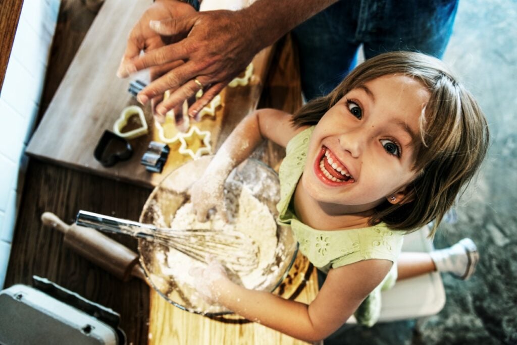 A young girl in a green dress smiles up at the camera while baking. Her hands are covered in flour, and a mixing bowl is in front of her. Cookie cutters and dough are on the table nearby, alongside an adult's hands rolling dough.