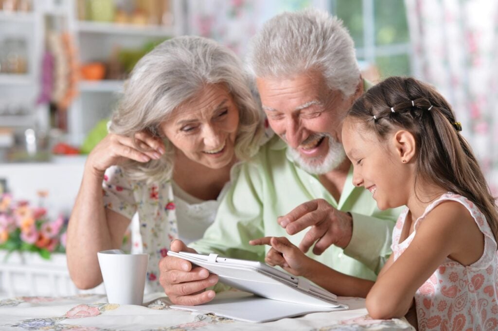 An elderly couple and a young girl sit at a table, smiling and looking at a tablet together. The scene is bright and cheerful, with a mug and floral decorations in the background.