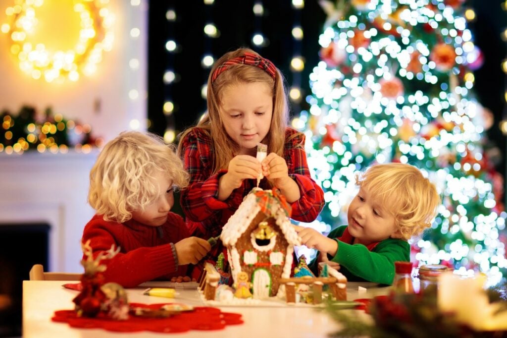 Three children decorate a gingerbread house at a table. A girl in a red headband is in the middle, with two boys on either side. The background features a brightly lit Christmas tree and festive decorations.