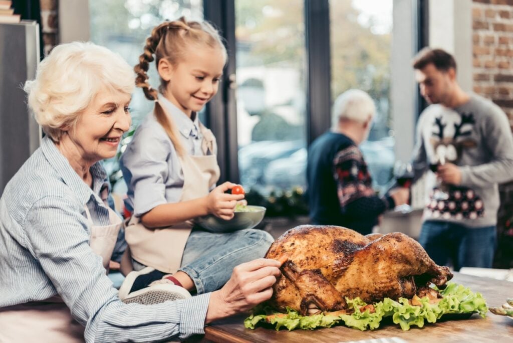 A grandmother and young girl are preparing a roasted turkey in a kitchen. The girl, sitting on the counter, holds a bowl with vegetables. In the background, two men in festive sweaters are talking near a window.