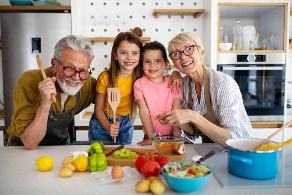 An elderly couple and two young girls smile while preparing a meal in a modern kitchen. The countertop has fresh vegetables, a blue pot, and kitchen utensils. The background features a refrigerator and an oven.