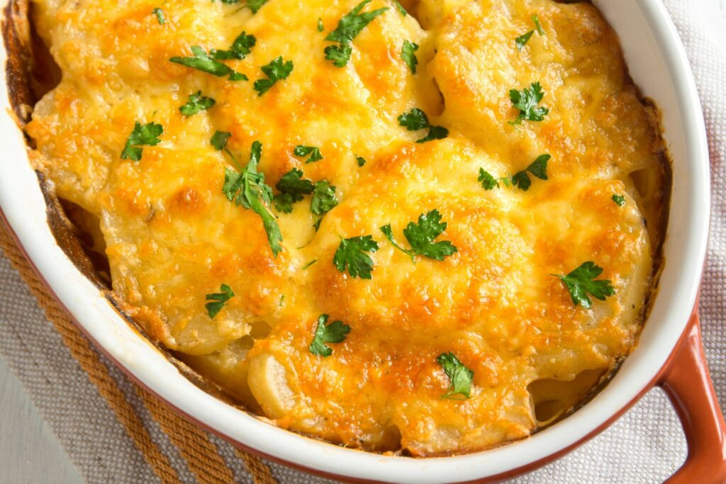 A close-up of a baked dish in an oval ceramic dish, featuring golden-brown scalloped potatoes topped with melted cheese and garnished with fresh parsley. The dish is placed on a cloth napkin.