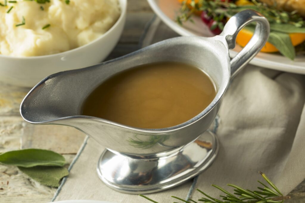 A silver gravy boat filled with brown gravy is placed on a table. In the background, a bowl of mashed potatoes and a plate with herbs and vegetables can be seen.