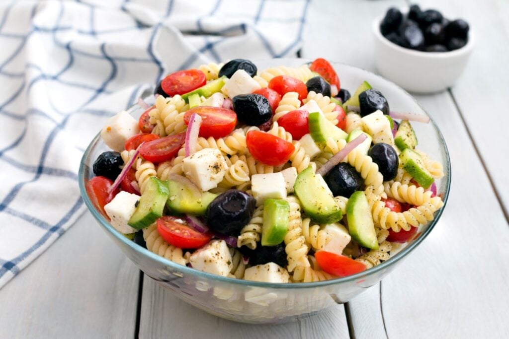 A glass bowl filled with pasta salad, including rotini, cherry tomatoes, black olives, cucumbers, mozzarella cheese cubes, and red onion slices. A small bowl of black olives is in the background, next to a checkered napkin.