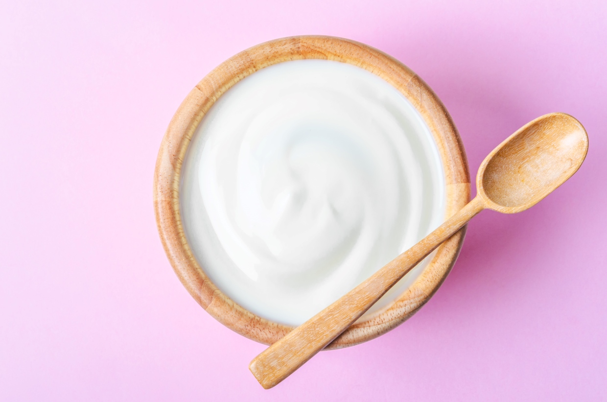 A wooden bowl filled with creamy white yogurt, accompanied by a wooden spoon resting on the bowl's edge, set against a pink background.