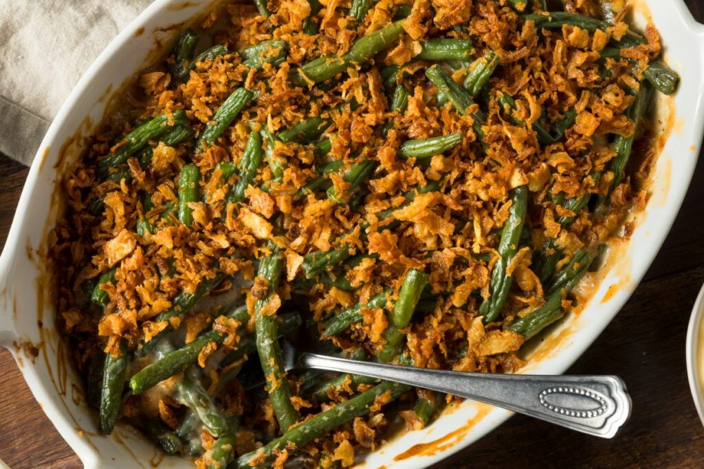 A baked dish of green bean casserole topped with crispy fried onions in an oval white casserole dish. A serving spoon is placed inside, ready to serve the dish. Rustic wooden table setting in the background.