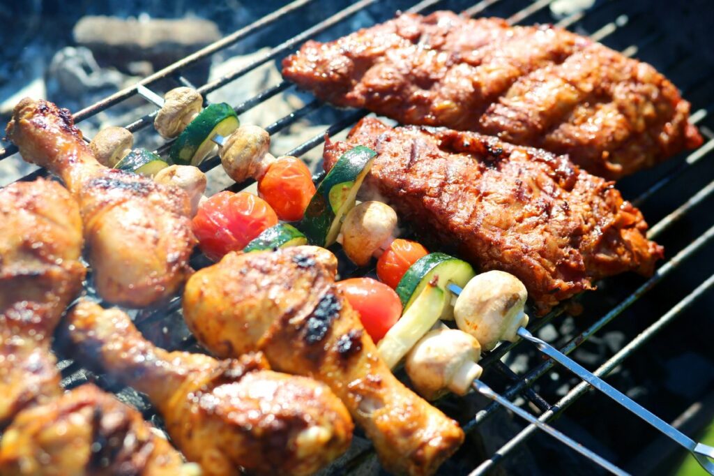 Grilled chicken drumsticks, vegetable skewers with mushrooms, tomatoes, and zucchini, and seasoned meat slices cooking over an open barbecue grill.