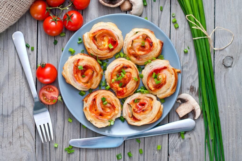 A plate of spiral puff pastries topped with chopped green onions and diced red peppers. Surrounding the plate are fresh tomatoes, mushrooms, and a bunch of green onions. A fork and knife are placed beside the plate on a wooden table.