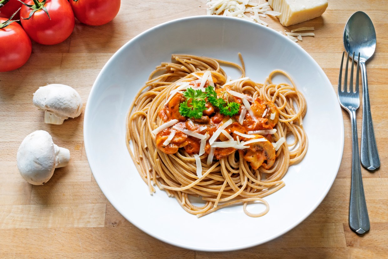 A plate of spaghetti topped with tomato sauce, shredded cheese, and parsley on a wooden table. Beside the plate are whole tomatoes, mushrooms, and a block of cheese. A fork and spoon are placed next to the plate.