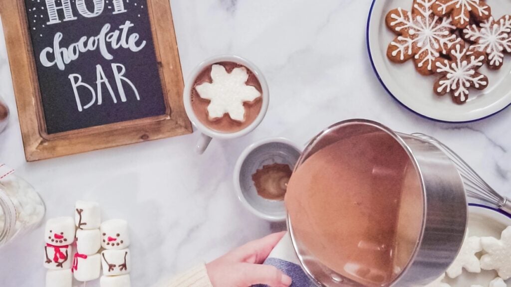 A person pours hot chocolate into a mug on a marble countertop. Nearby are decorated cookies, a sign reading "Hot Chocolate Bar," and marshmallows with snowman faces.