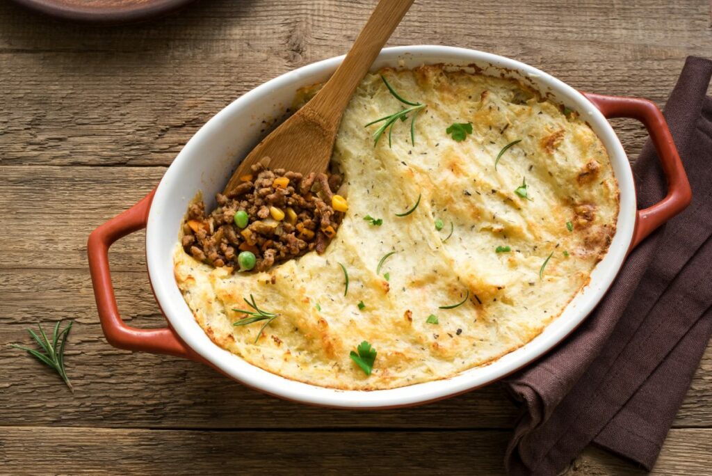 A baked shepherd's pie in an oval dish with mashed potato topping. A wooden spoon reveals the filling of ground meat, peas, and corn. Garnished with rosemary and parsley. Dish rests on a wooden surface with a brown cloth beside it.