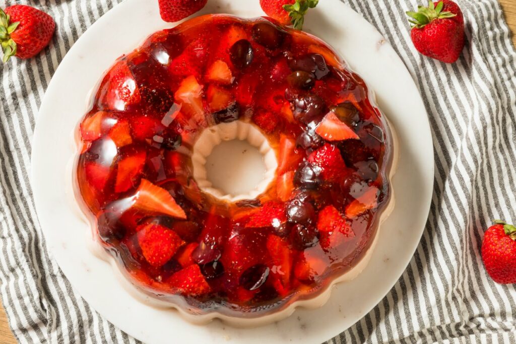 A round fruit jelly dessert on a white plate, featuring assorted fruits like strawberries and cherries. It sits on a striped cloth with fresh strawberries scattered around.