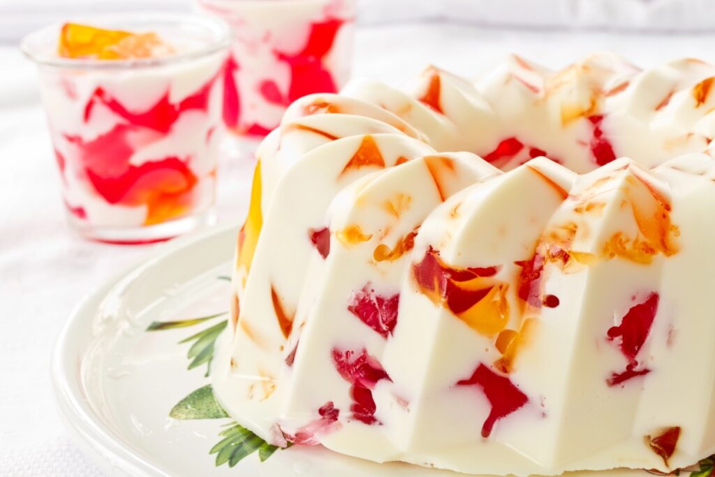 A vibrant gelatin mold dessert with red and orange jelly pieces embedded in creamy white gelatin, presented on a decorative plate. In the background, small cups hold similar servings.