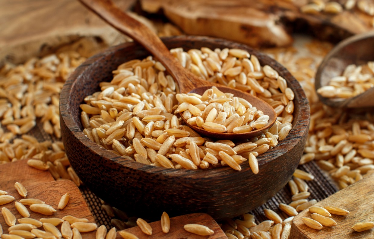 A wooden bowl filled with uncooked orzo pasta, with a wooden spoon resting inside. More orzo is scattered around the bowl on a rustic wooden surface, creating a warm and earthy ambiance.