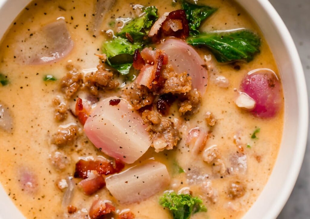 A close-up of a creamy soup with chunks of radishes, sausage, crispy bacon, and kale, seasoned with black pepper. The soup has a rich broth and is garnished with fresh greens.