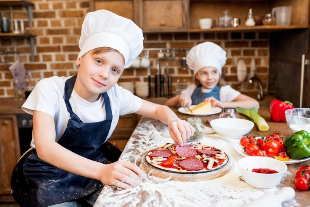 Two children in chef hats and aprons are in a kitchen preparing pizzas. One is adding toppings to a pizza, while the other is grating cheese. Fresh vegetables and ingredients are visible on the table.