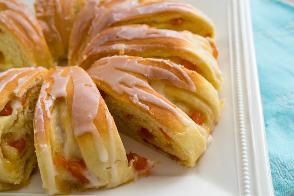 A close-up of a sliced, glazed pastry ring on a white platter. The bread is golden brown with a visible layer of icing on top. Small pieces of orange or apricot filling peek out from between the layers.