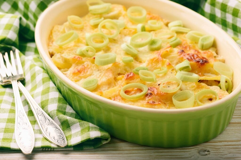 A green casserole dish filled with baked gratin, topped with sliced leeks. Two ornate silver forks are placed beside the dish on a green and white checkered cloth.