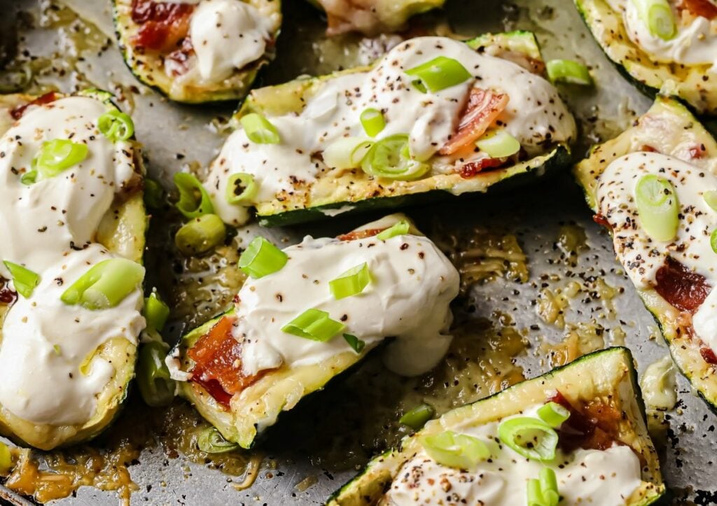 Stuffed zucchini boats topped with melted cheese, sour cream, chopped green onions, and black pepper. The zucchini boats are arranged on a baking tray, with a crispy, cheesy texture visible on the edges.