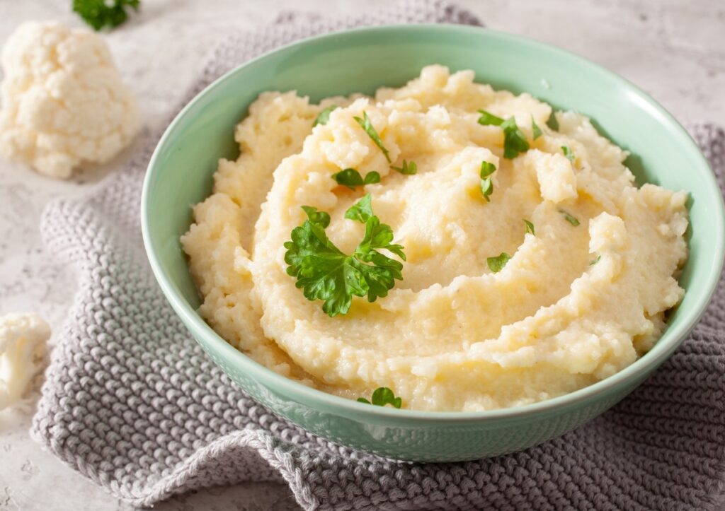 A bowl of creamy mashed cauliflower garnished with fresh parsley sits on a folded gray cloth. The table is speckled, and a small piece of cauliflower is visible in the background.