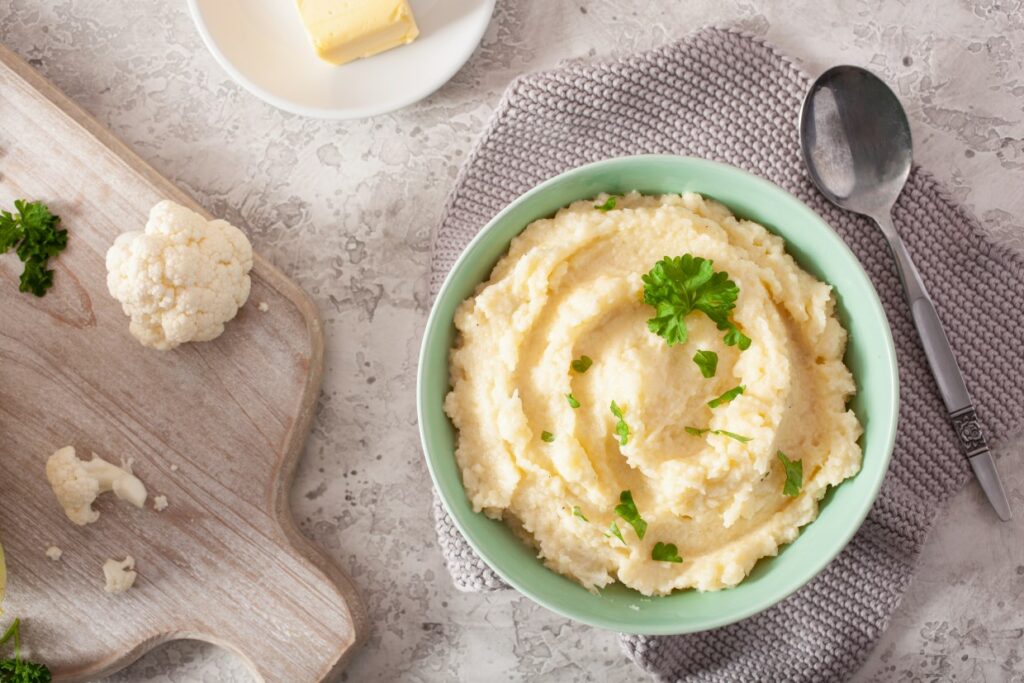 A bowl of creamy mashed cauliflower garnished with parsley on a textured napkin. A spoon is beside it. Nearby, a cutting board with cauliflower florets and a butter dish on a gray surface.