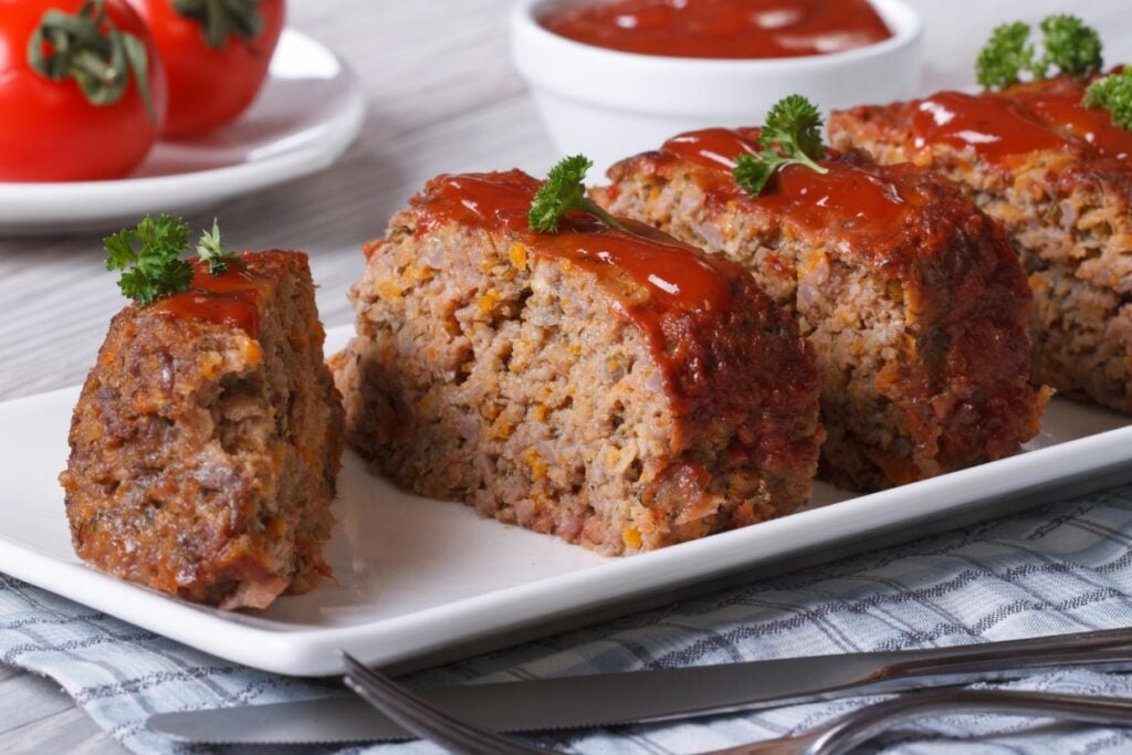 Slices of glazed meatloaf garnished with parsley on a white plate, with a bowl of sauce in the background. Fresh tomatoes are seen in the top left corner.