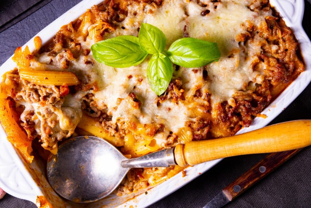 A baked pasta casserole topped with melted cheese and garnished with fresh basil leaves. A wooden-handled spoon rests inside the casserole dish, ready to serve. The scene conveys a warm, inviting meal setting.