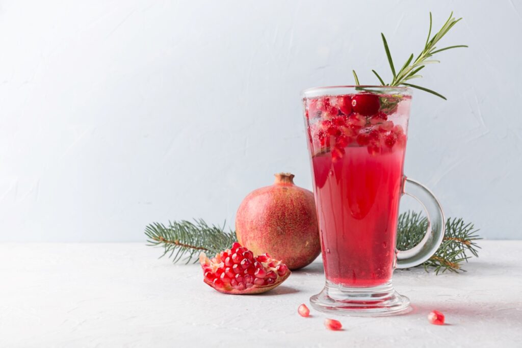 A tall glass of pomegranate drink garnished with rosemary sits on a white surface. Next to it, a whole pomegranate and a cut half revealing seeds are displayed. Pine branches add a festive touch to the scene.