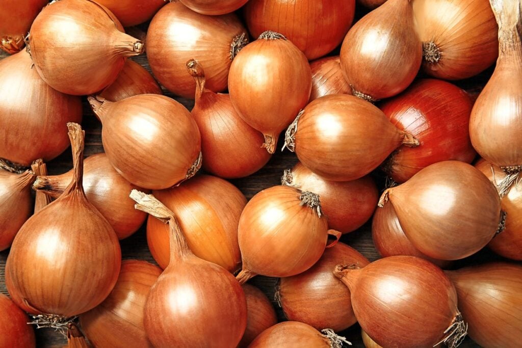 A heap of fresh brown onions with papery skin, closely packed together. The onions' round shapes and earthy tones are captured in natural light, highlighting their texture and slight sheen.