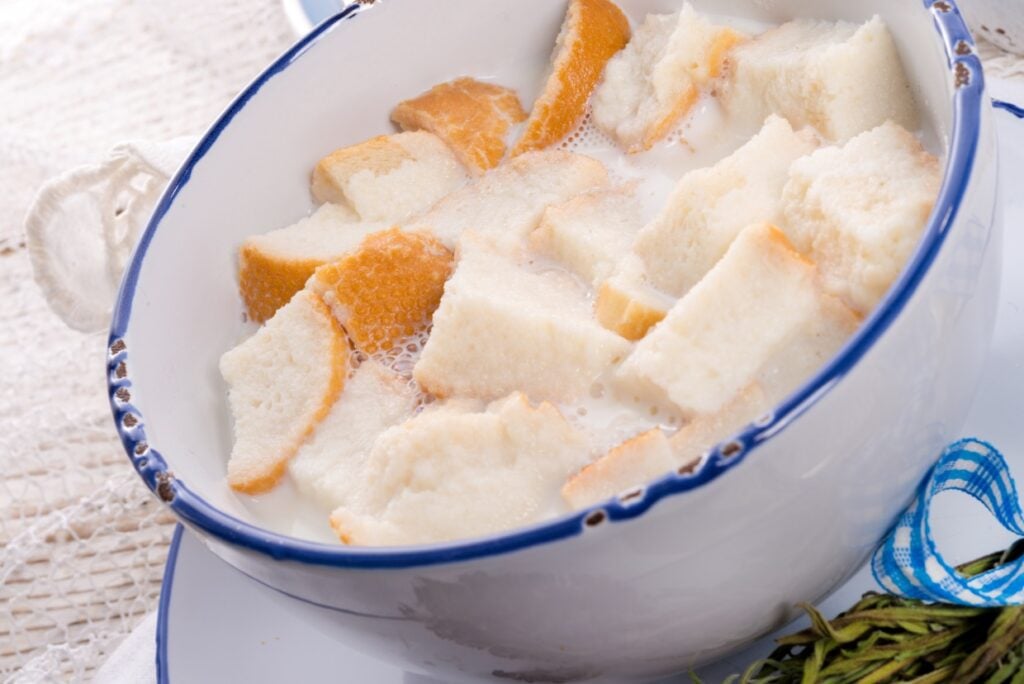 A bowl filled with chunks of bread soaked in milk, displayed on a white tablecloth. The bread has a mix of crust and soft pieces, and there is a blue-rimmed edge on the bowl. Fresh herbs are partially visible beside the bowl.