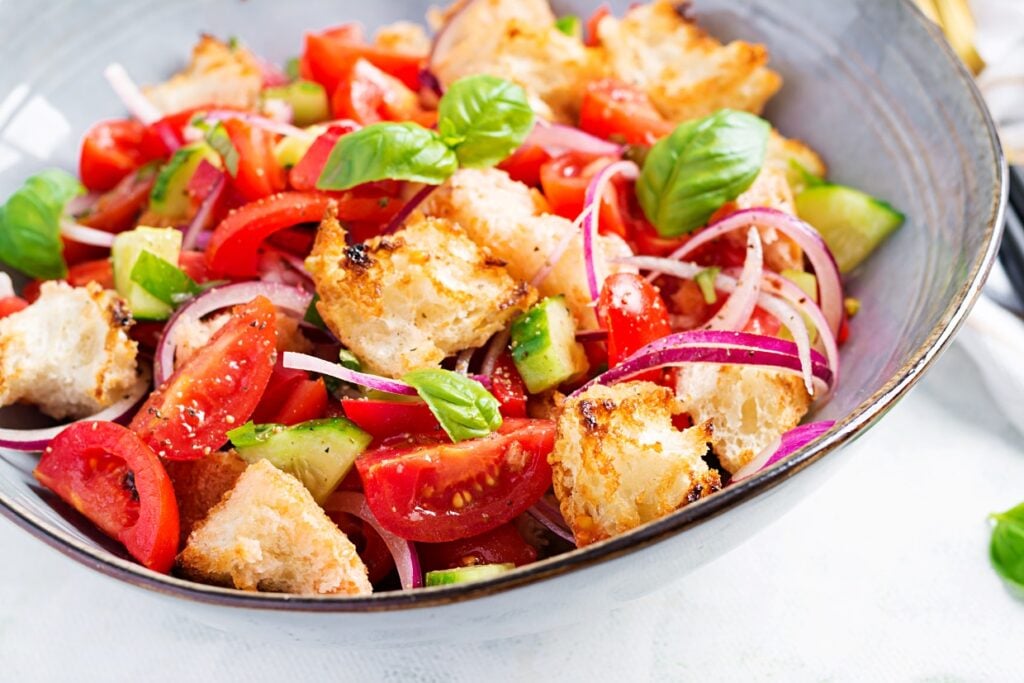 A colorful salad in a bowl featuring diced tomatoes, cucumbers, red onion slices, crispy croutons, and fresh basil leaves. The ingredients are vibrant and fresh, presented in a gray bowl on a light-colored surface.