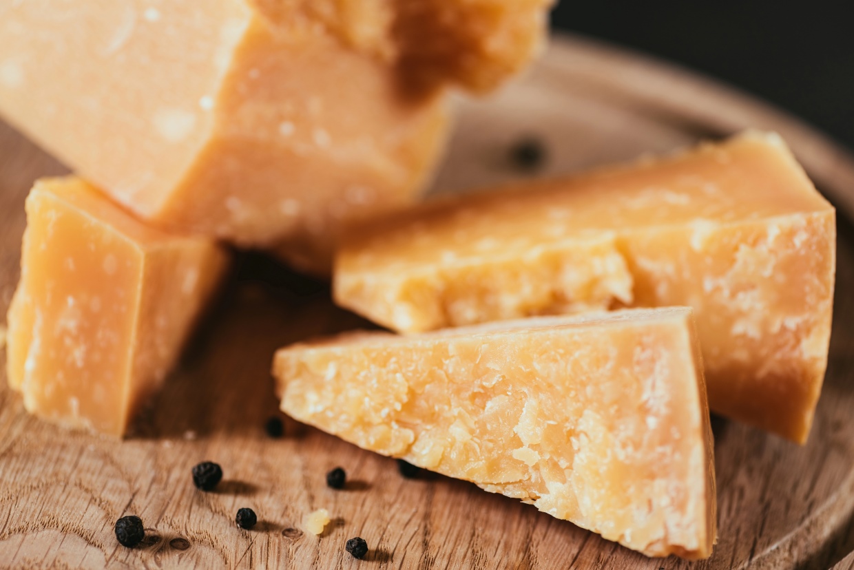 Close-up of several wedge-shaped pieces of hard cheese on a wooden surface, with scattered black peppercorns around them. The cheese has a textured, crumbly appearance.