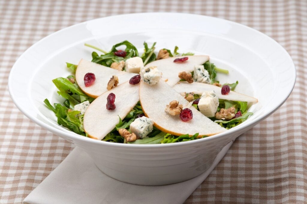 A white bowl filled with a fresh salad containing arugula, pear slices, walnuts, blue cheese, and dried cranberries, placed on a folded napkin. The background is a checkered tablecloth.